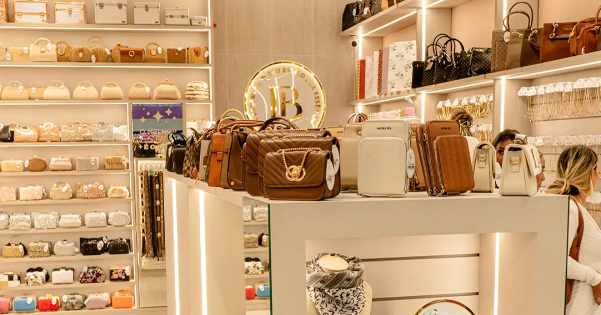 Loja Donna Bendita com prateleiras iluminadas exibindo bolsas e acessórios femininos variados, desde clutches a bolsas estruturadas, em ambiente sofisticado de shopping.